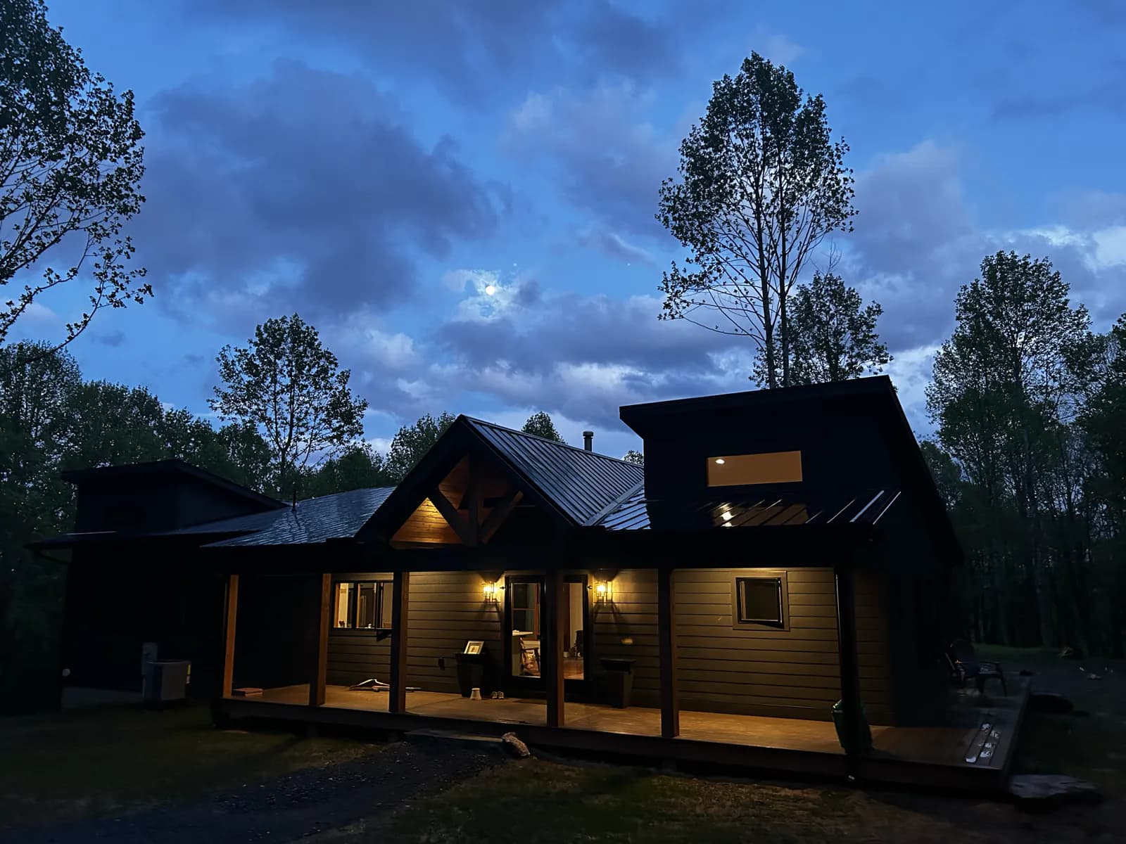 The finished home at blue hour in April, porch lights glowing under the timber-frame gable and the surrounding trees leafed out for spring