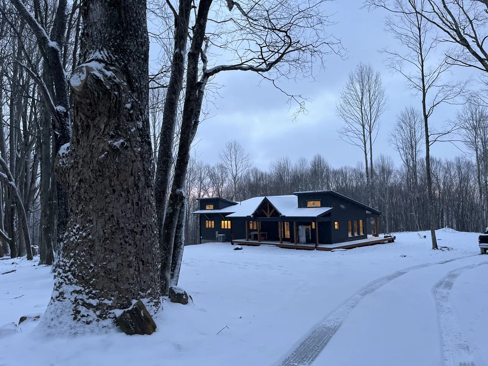 The same home at dusk in March, windows lit warm against the snow, with tire tracks curving up the drive past a snow-covered maple